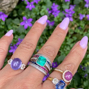 May include: A hand displays five gemstone rings. The rings feature a variety of stones, including a large emerald, a purple stone, a pink stone, and a blue stone. The rings are set in gold and silver bands, with diamond accents.