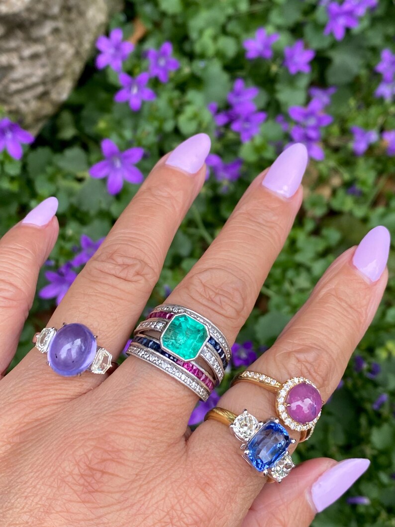 May include: A hand displays five gemstone rings. The rings feature a variety of stones, including a large emerald, a purple stone, a pink stone, and a blue stone. The rings are set in gold and silver bands, with diamond accents.