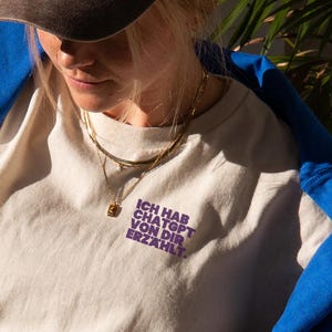 Puede incluir: Una persona con una camiseta beige con texto morado, una sudadera azul y una gorra marr&oacute;n. La persona est&aacute; sentada en una mesa con una bebida de caf&eacute;, un cruas&aacute;n y un libro. El texto de la camiseta dice "ICH HAB CHATGPT VON DIR ERZ&Auml;HLT."