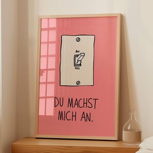 May include: A framed art print featuring a pink background with a light switch illustration. The switch is labeled "AN" and "AUS". Below the switch, the text reads "DU MACHST MICH AN."