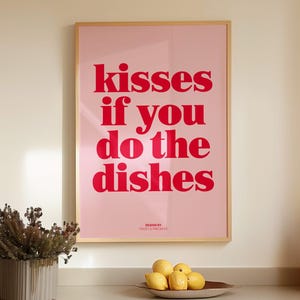 May include: A framed pink poster with the text "kisses if you do the dishes" in red. The poster is hanging above a wooden countertop with a decorative vase, a bowl of lemons, and brown bottles.