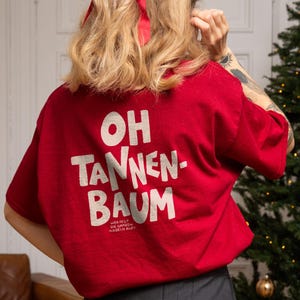May include: A red short-sleeved t-shirt featuring the white text OH TANNEN-BAUM and smaller text below it, paired with a dark grey pleated skirt. The shirt is worn with a red hair ribbon, and the overall ensemble is captured from the back.
