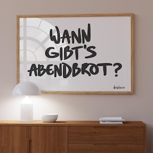May include: A framed print with the text "WANN GIBT'S ABENDBROT?" in black lettering hangs above a wooden cabinet. A white lamp and bowl sit on the cabinet. The cabinet has two doors and two drawers.