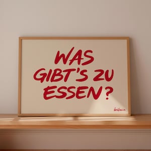 May include: A framed print with the text "WAS GIBT'S ZU ESSEN?" in red, handwritten-style lettering. The print is set against a cream background and displayed on a wooden bench.