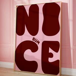 Może przedstawiać: Oprawiony wydruk artystyczny z napisem "NICE" w dużych, ciemnoczerwonych literach bąbelkowych, z napisem "BUM" w mniejszych literach i słowem "CE" poniżej. Tło jest jasnoróżowe, a rama złota.