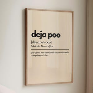 May include: A framed print with the words "deja poo" in a bold, black font. Below the title is the phonetic pronunciation and a German definition. The print is in a light wood frame and hangs on a white wall.