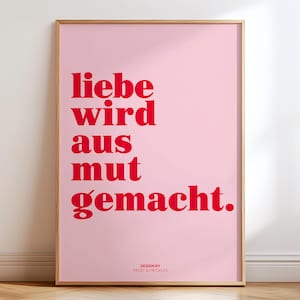 May include: A framed pink art print with red text that reads "liebe wird aus mut gemacht." The print is in a wooden frame and is displayed against a white wall. The bottom right corner of the print reads "DESIGN BY FROST & FRECKLER."