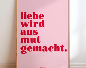 May include: A framed pink art print with red text that reads "liebe wird aus mut gemacht." The print is in a wooden frame and is displayed against a white wall. The bottom right corner of the print reads "DESIGN BY FROST & FRECKLER."
