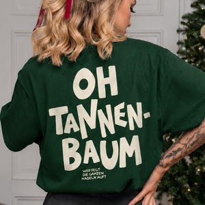 May include: A dark green short-sleeved t-shirt featuring the cream-coloured text OH TANNEN-BAUM and smaller text below reading WER FEGT DIE GANZEN NADELN AUF? paired with a charcoal grey pleated skirt and black patterned tights.