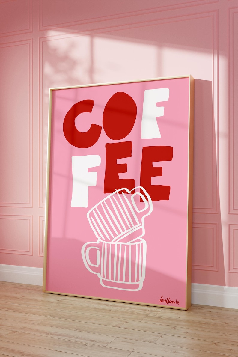 May include: A framed pink art print with the word "COFFEE" in bold red and white letters. Below the text are two stacked coffee mugs with white vertical stripes. The artwork is displayed against a pink wall.