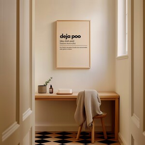 May include: A framed print with the word "deja poo" in black text. The print is hanging above a wooden desk with a small chair and a blanket draped over it. A small plant and soap dispenser are on the desk.