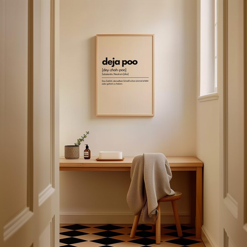 May include: A framed print with the word "deja poo" in black text. The print is hanging above a wooden desk with a small chair and a blanket draped over it. A small plant and soap dispenser are on the desk.