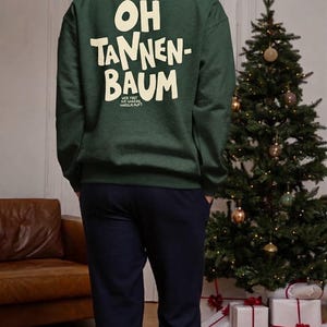 May include: A dark green long-sleeve crewneck sweatshirt featuring the cream-colored text "OH TANNEN-BAUM" printed on the back, with smaller text underneath that reads "WER FEET DIE GANSEN NASSEN PUPT". The sweatshirt has a ribbed collar and cuffs.