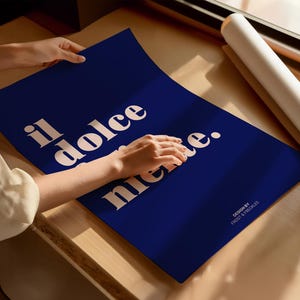 May include: A rectangular dark blue poster featuring the cream-colored text "il dolce far niente." in a bold serif font. The bottom right corner includes the smaller text "DESIGN BY FROST & FRECKLES." The poster is displayed on a flat surface.