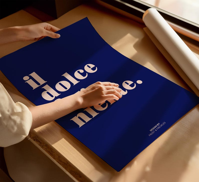 May include: A rectangular dark blue poster featuring the cream-colored text "il dolce far niente." in a bold serif font. The bottom right corner includes the smaller text "DESIGN BY FROST & FRECKLES." The poster is displayed on a flat surface.