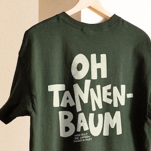 May include: A dark forest green short-sleeved t-shirt featuring the large white text "OH TANNEN-BAUM" and smaller text below reading "WER FEGT DIE GANZEN NADELN AUF?" The shirt is displayed on a wooden hanger against a neutral background.