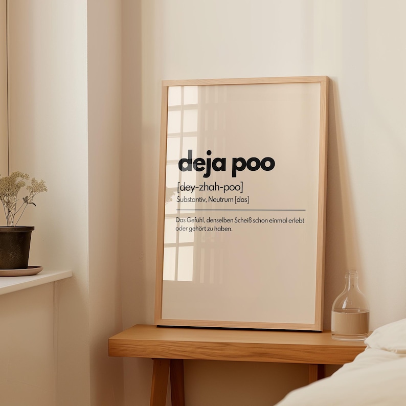 May include: A framed print with the words "deja poo" in a bold, black font. Below the title is the phonetic pronunciation and the word "Substantiv, Neutrum [das]" in a smaller font. The print is on a light beige background.