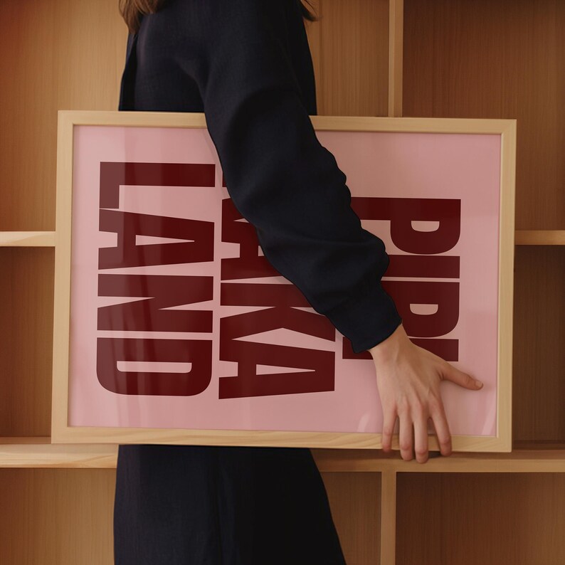 Peut inclure: Une affiche encadr&eacute;e avec les mots "PIKA LAND" en lettres majuscules rouge fonc&eacute; sur un fond rose clair. Le cadre est en bois clair. L'&oelig;uvre est tenue par une personne v&ecirc;tue d'un haut noir.
