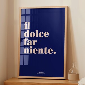 May include: A rectangular wall art print in a light wood frame featuring a deep blue background with the white text "il dolce far niente." and the smaller text "DESIGN BY FROST & FRECKLES" at the bottom, displayed on a wooden surface.