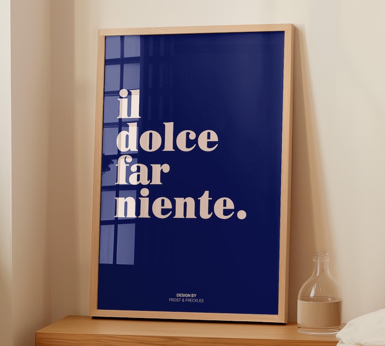May include: A rectangular wall art print in a light wood frame featuring a deep blue background with the white text "il dolce far niente." and the smaller text "DESIGN BY FROST & FRECKLES" at the bottom, displayed on a wooden surface.