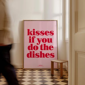 May include: A framed pink poster with the text "kisses if you do the dishes" in red. The poster is in a gold frame and is leaning against a white wall. A small wooden stool sits in front of the poster.