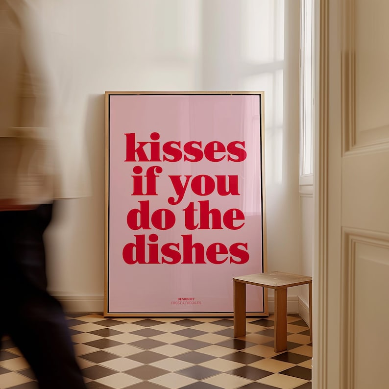 May include: A framed pink poster with the text "kisses if you do the dishes" in red. The poster is in a gold frame and is leaning against a white wall. A small wooden stool sits in front of the poster.