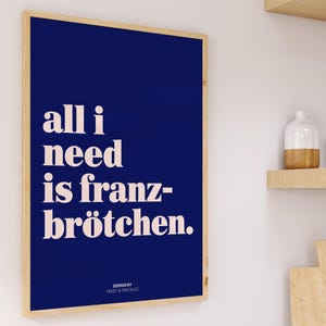 May include: A rectangular wall art print in a light wood frame featuring a deep blue background with the white text "all i need is franz-br&ouml;tchen." and the smaller text "DESIGN BY FROST & FRECKLES" positioned at the bottom center of the print.
