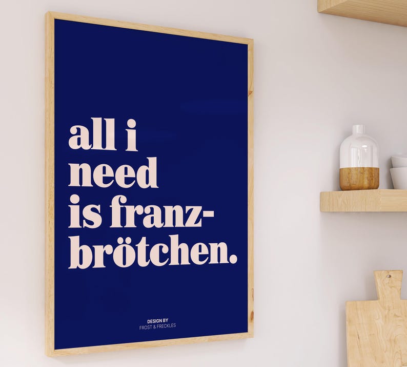 May include: A rectangular wall art print in a light wood frame featuring a deep blue background with the white text "all i need is franz-br&ouml;tchen." and the smaller text "DESIGN BY FROST & FRECKLES" positioned at the bottom center of the print.