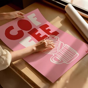 May include: A pink poster with the word "COFFEE" in large red and white letters, along with a graphic of two coffee mugs. The poster is being held by a person. A rolled-up poster is in the background.