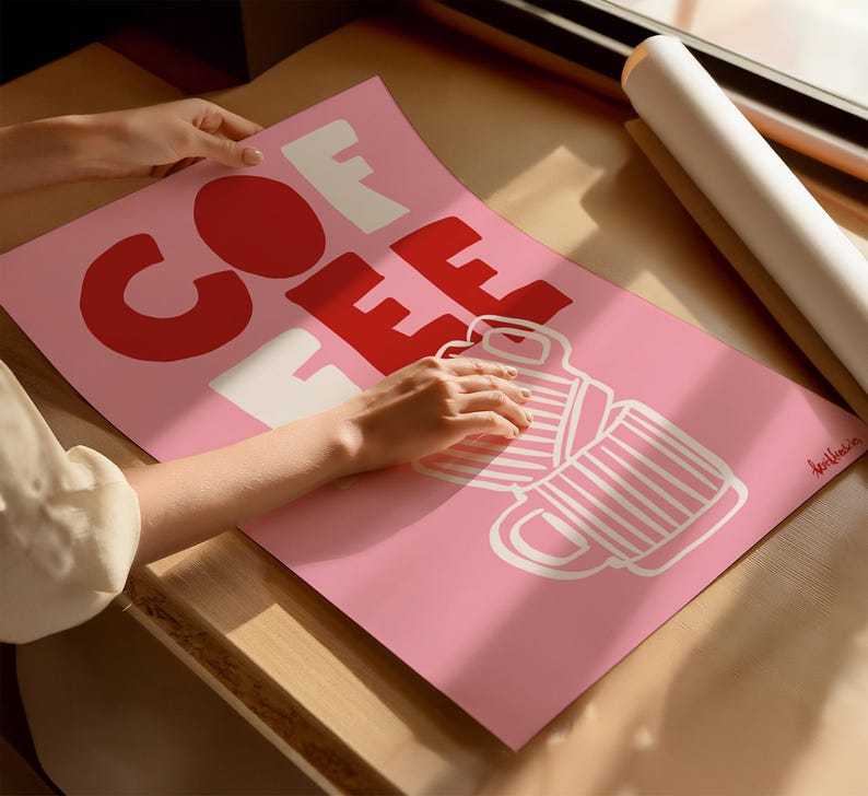 May include: A pink poster with the word "COFFEE" in large red and white letters, along with a graphic of two coffee mugs. The poster is being held by a person. A rolled-up poster is in the background.
