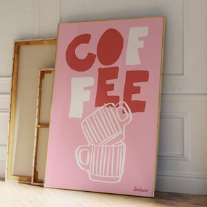 May include: A pink framed art print with the word "COFFEE" in red and white block letters. Two stacked coffee mugs are drawn in white. The artwork is leaning against a white wall and a wooden framed canvas.
