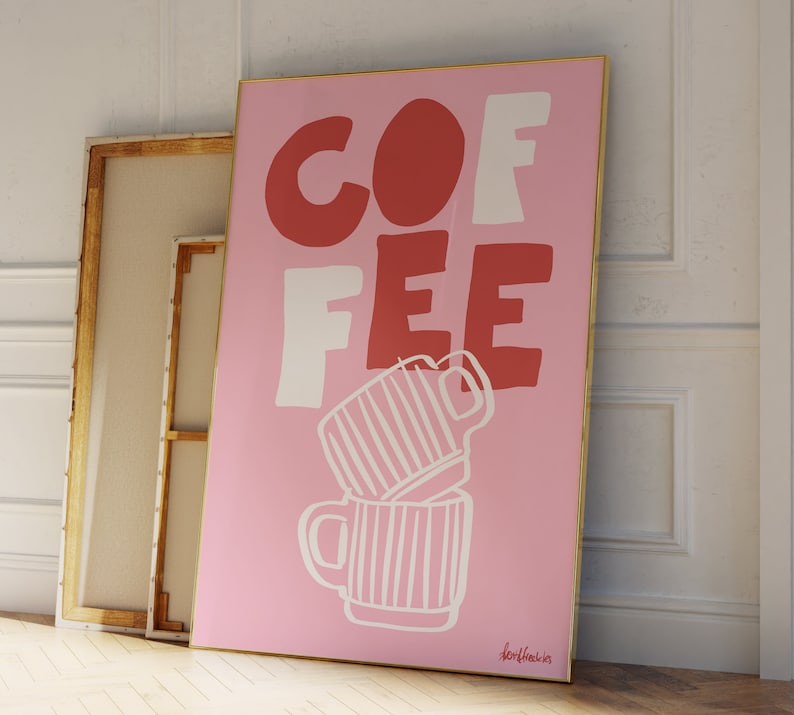 May include: A pink framed art print with the word "COFFEE" in red and white block letters. Two stacked coffee mugs are drawn in white. The artwork is leaning against a white wall and a wooden framed canvas.