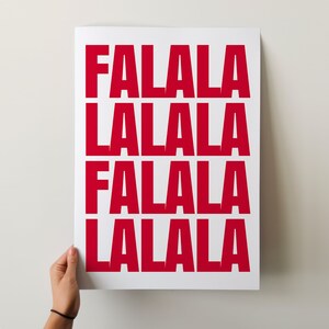 May include: A white poster with the words "FALALA LALALA FALALA LALALA" printed in bold, red letters. The poster is held by a person's hand against a white background. The design is simple and festive.