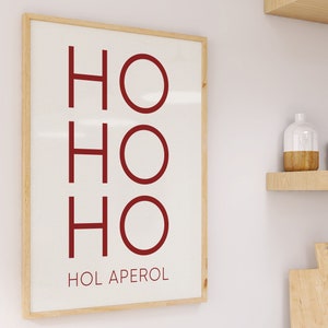 May include: A rectangular wall art print in a light wood frame featuring the red text HO HO HO followed by the text HOL APEROL in a smaller font on a white background. The design is displayed vertically with clean lines and a simple layout.