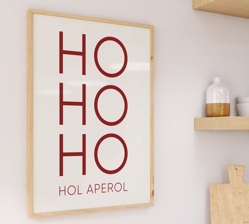 May include: A rectangular wall art print in a light wood frame featuring the red text HO HO HO followed by the text HOL APEROL in a smaller font on a white background. The design is displayed vertically with clean lines and a simple layout.