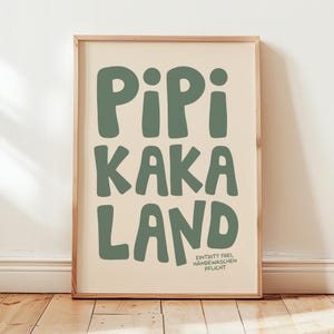 May include: A framed wall art print featuring the text PIPI KAKA LAND in large, bold, sage green letters against a cream background. In the bottom right corner, smaller text reads EINTRITT FREI, HÄNDEWASCHEN PFLICHT in the same sage green color.