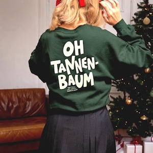 May include: A dark green long-sleeve sweatshirt featuring the white text "OH TANNEN-BAUM" printed on the back. The garment has a crew neckline and a relaxed fit, paired with a dark-colored pleated skirt. A red ribbon is tied in the hair.