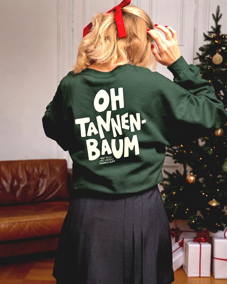 May include: A dark green long-sleeve sweatshirt featuring the white text "OH TANNEN-BAUM" printed on the back. The garment has a crew neckline and a relaxed fit, paired with a dark-colored pleated skirt. A red ribbon is tied in the hair.