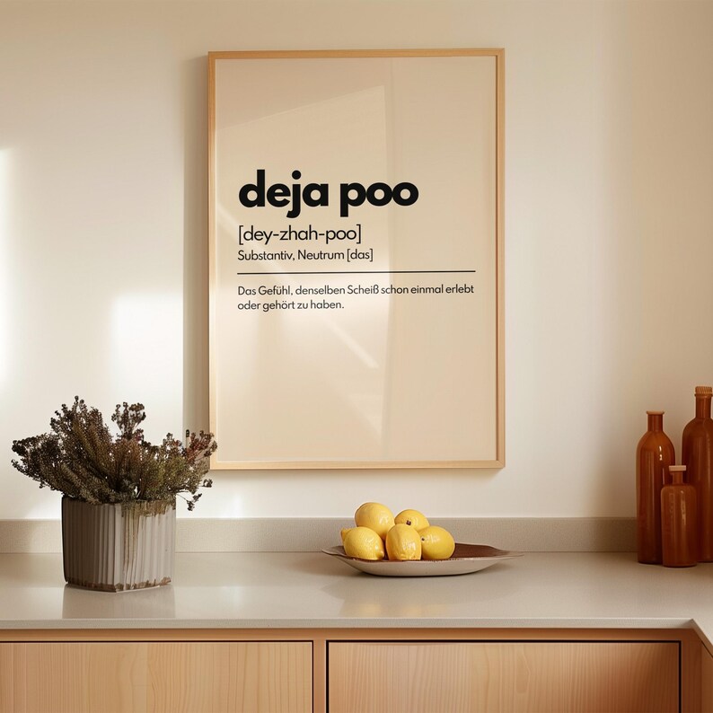 May include: A framed print with the text "deja poo" in black, with phonetic pronunciation and German definition. The print is displayed above a countertop with a plant, lemons, and decorative bottles.