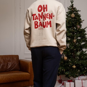 May include: A beige long-sleeve sweatshirt featuring the red text OH TANNEN-BAUM printed on the back. Below the main text, smaller red text reads WER FEGT DIE GANZEN NADELN AUF. The sweatshirt has a crew neckline and ribbed cuffs and hem.