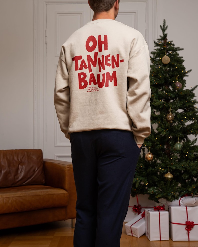 May include: A beige long-sleeve sweatshirt featuring the red text OH TANNEN-BAUM printed on the back. Below the main text, smaller red text reads WER FEGT DIE GANZEN NADELN AUF. The sweatshirt has a crew neckline and ribbed cuffs and hem.