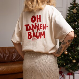 May include: An off-white short-sleeve t-shirt featuring red text on the back that reads OH TANNEN-BAUM and smaller text below that reads WER FEGT DIE GANZEN NADELN AUF? The shirt is paired with a silver sequined skirt that has a textured, shimmering finish.