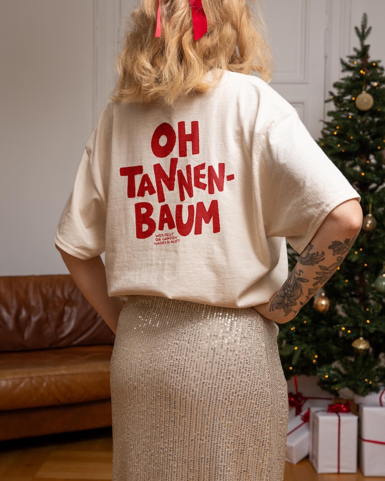 May include: An off-white short-sleeve t-shirt featuring red text on the back that reads OH TANNEN-BAUM and smaller text below that reads WER FEGT DIE GANZEN NADELN AUF? The shirt is paired with a silver sequined skirt that has a textured, shimmering finish.