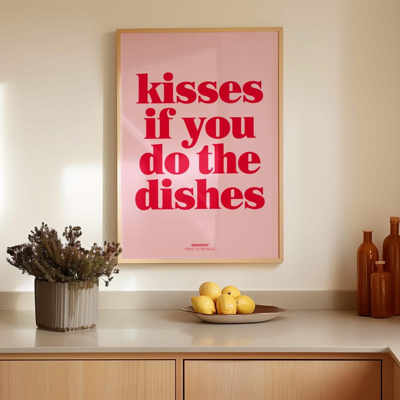 May include: A framed pink poster with the text "kisses if you do the dishes" in red. The poster is hanging above a wooden countertop with a decorative vase, a bowl of lemons, and brown bottles.