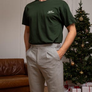 May include: A dark green short-sleeved t-shirt featuring the white text MERRY CHRISTMAS embroidered on the left chest, paired with light grey trousers that have a pleated front and side buckle adjusters at the waistband.
