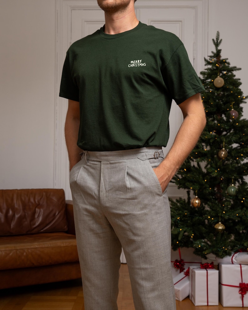 May include: A dark green short-sleeved t-shirt featuring the white text MERRY CHRISTMAS embroidered on the left chest, paired with light grey trousers that have a pleated front and side buckle adjusters at the waistband.