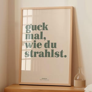 May include: A framed art print with the text "guck mal, wie du strahlst" in green lettering. The print has a light beige background and a natural wood frame. The words translate to "look how you shine".