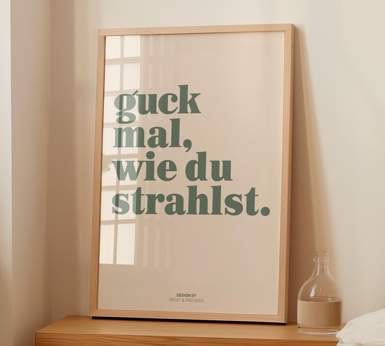May include: A framed art print with the text "guck mal, wie du strahlst" in green lettering. The print has a light beige background and a natural wood frame. The words translate to "look how you shine".