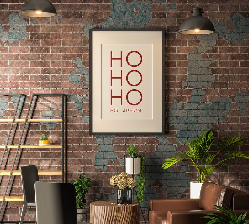 May include: A rectangular wall art print in a black frame featuring the red text HO HO HO and HOL APEROL in a sans-serif font on a cream background. The print is centered on a rustic brick wall with visible mortar and weathered texture.