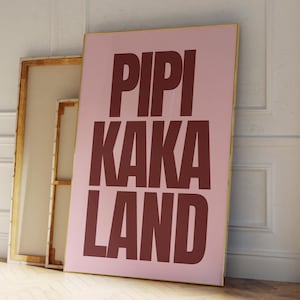 Peut inclure: Une affiche rose encadr&eacute;e avec les mots "PIPI KAKA LAND" en grandes lettres majuscules rouge fonc&eacute;. L'affiche est appuy&eacute;e contre un mur blanc et une toile encadr&eacute;e en bois.